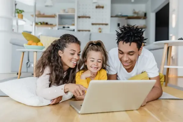 A smiling family of three, including a mother, father, and young daughter, is lying on the floor looking at a laptop together.